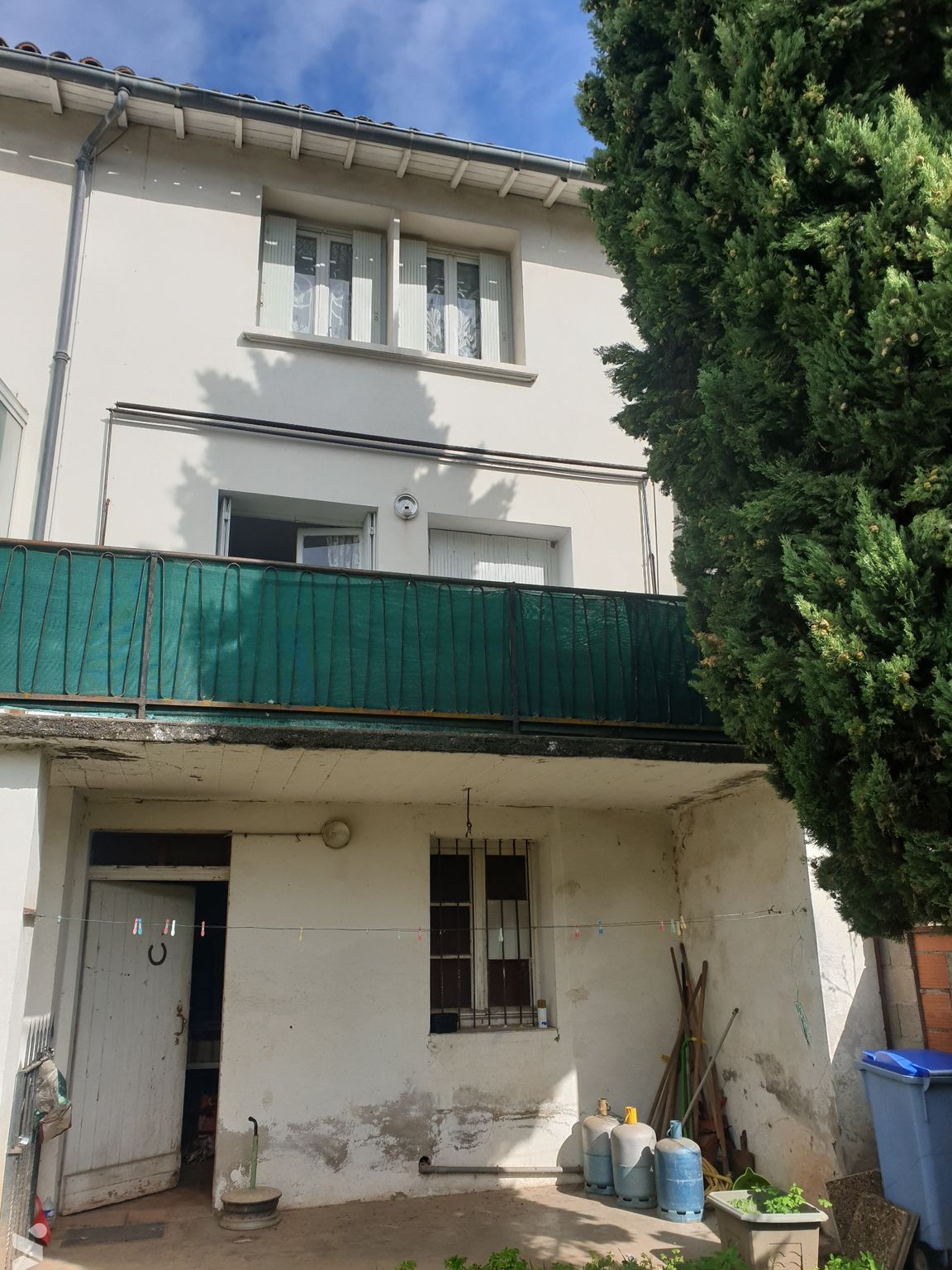 Bâtiment blanc de deux étages avec balcon vert, porte ouverte, poubelle bleue et grand arbre vert.