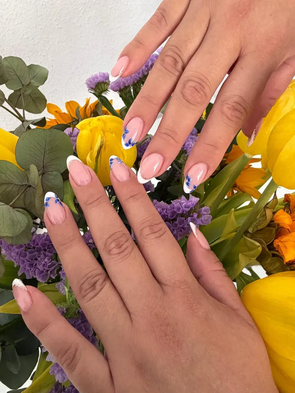 Manos con uñas pintadas de rosa con la punta francesa, descansando sobre un ramo de flores amarillas y moradas.