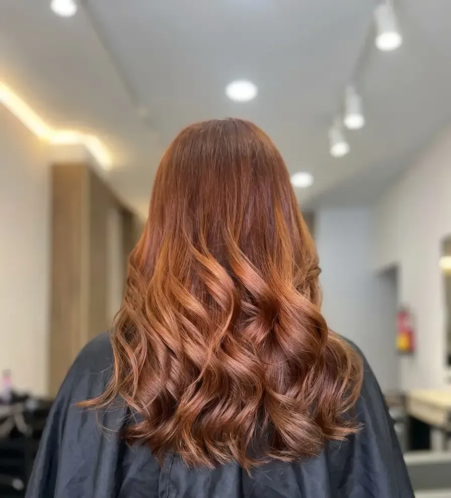 Vista posterior de una larga melena cobriza ondulada en una silla de peluquería, con puntas rizadas y reflejos cálidos.