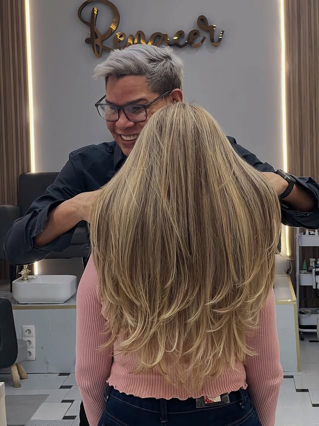 Peluquera arreglando el largo cabello rubio de una clienta en un salón de belleza, con un espejo y un letrero al fondo.