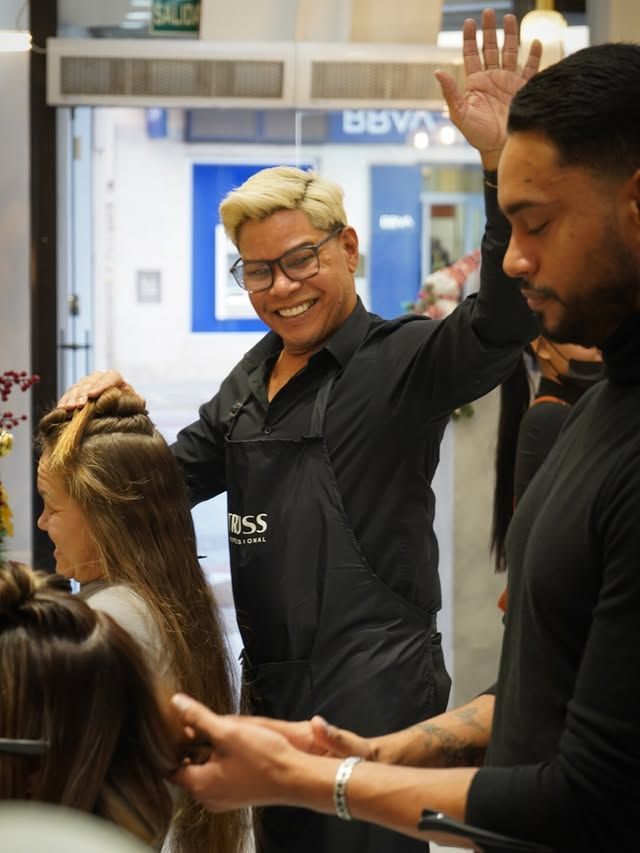 Una peluquera sonríe y saluda en una peluquería mientras sus clientes esperan cerca.