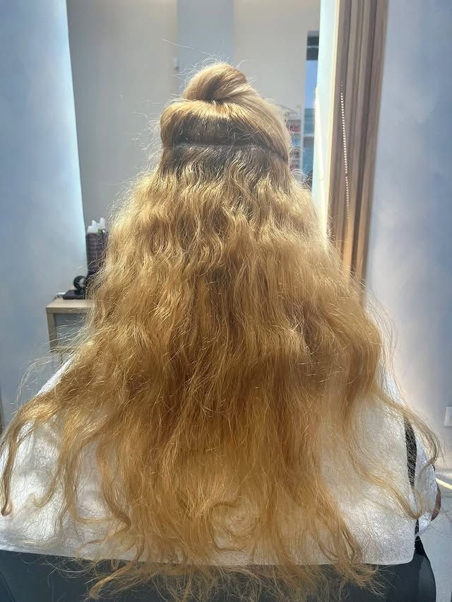 Vista trasera de una persona con cabello rubio largo y ondulado recogido a medias en una silla de peluquería.