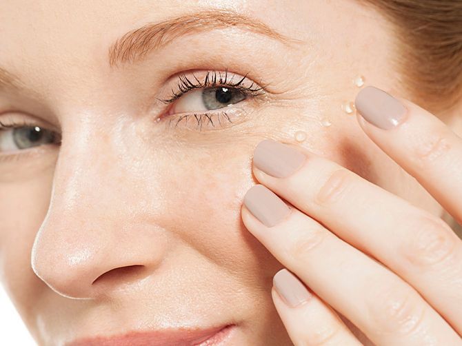 Primer plano de un rostro sonriente con una mano aplicándose crema en la mejilla cerca del ojo.