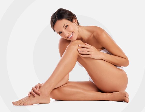 Mujer sentada sobre un fondo blanco, sonriendo y tocándose la pierna en una pose de estudio.