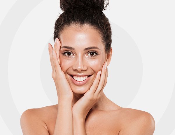 Mujer sonriente con el pelo recogido en un moño, tocándose suavemente la cara contra un fondo claro.