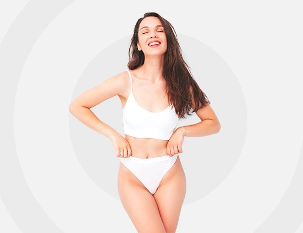 Mujer en ropa interior blanca posando sobre un fondo gris claro, sonriendo con las manos en las caderas.