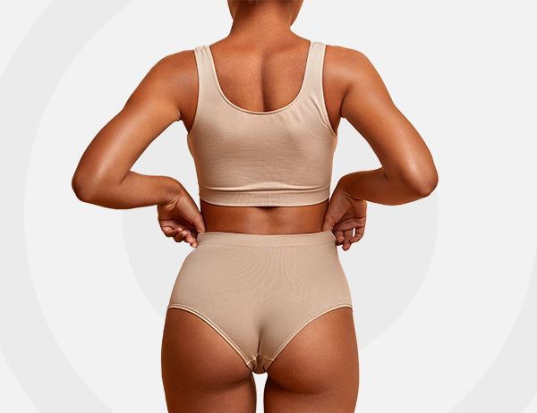 Mujer con sujetador deportivo y bragas color beige ajustándose la cinturilla, vista de espaldas sobre un fondo claro.