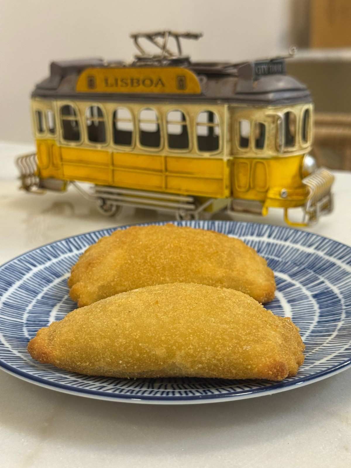Dos buñuelos dorados sobre un plato azul estampado con un tranvía amarillo de Lisboa al fondo.