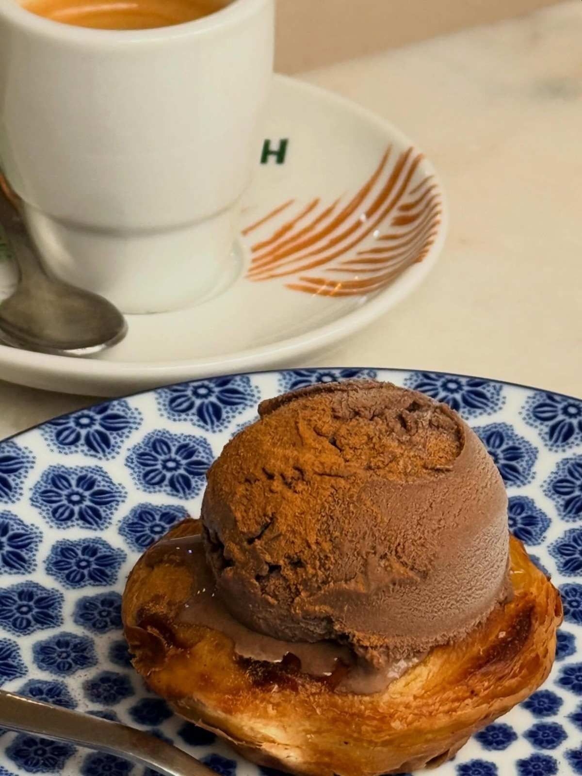Plato con estampado azul con un pastelito cubierto de helado de chocolate junto a una taza de café.