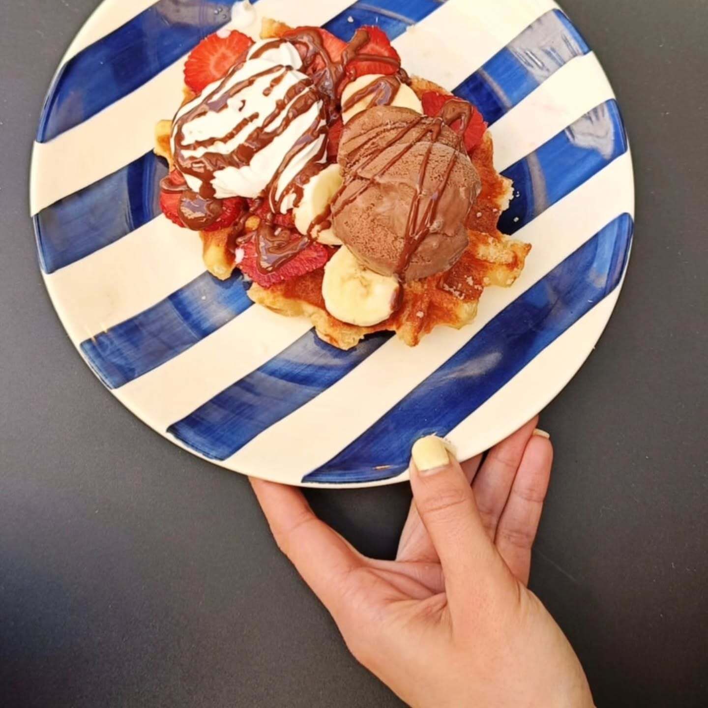 Una mano sostiene un plato a rayas azules y blancas con un postre cubierto con sirope de chocolate y crema batida.