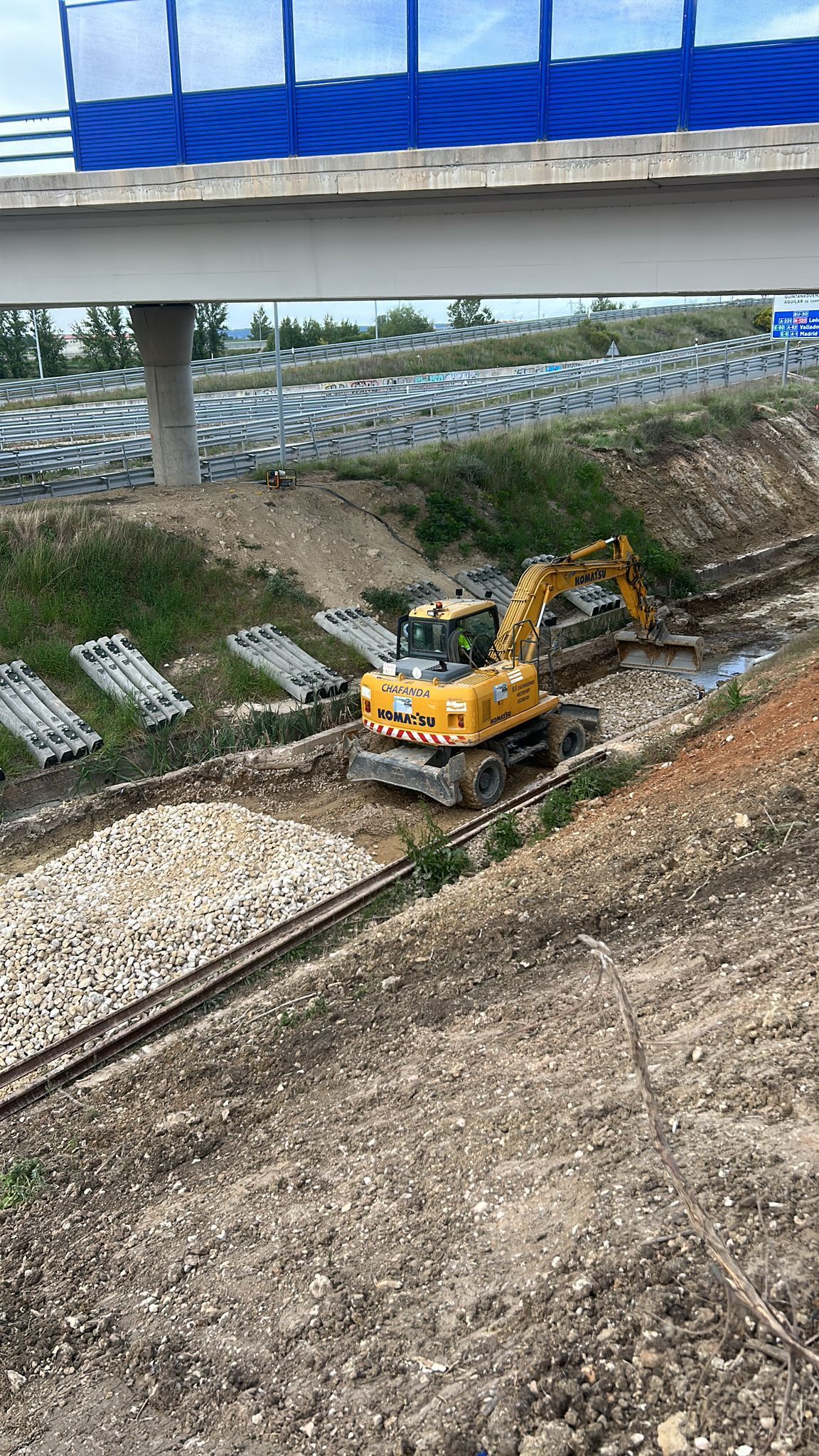 Una excavadora amarilla excava en un terraplén inclinado debajo de un puente.