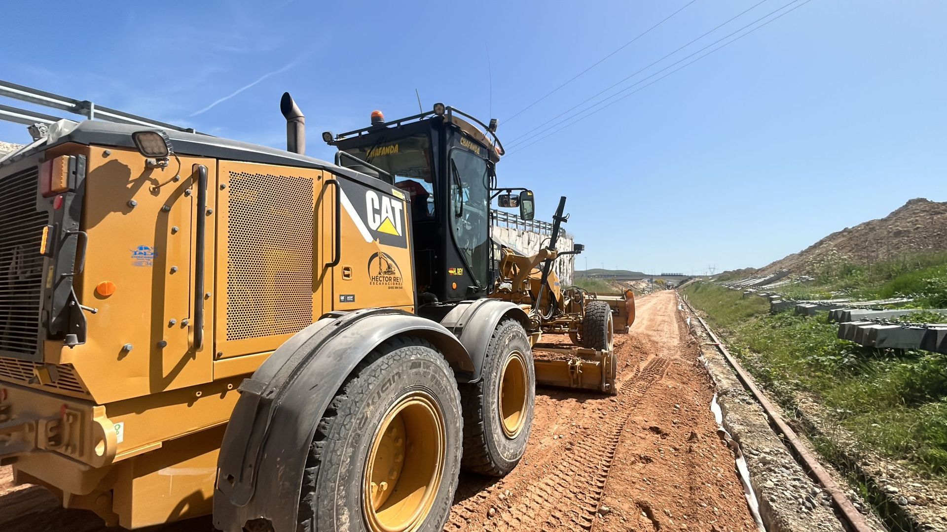 Niveladoras amarillas trabajando en un camino de tierra junto a campos verdes bajo un cielo azul despejado.