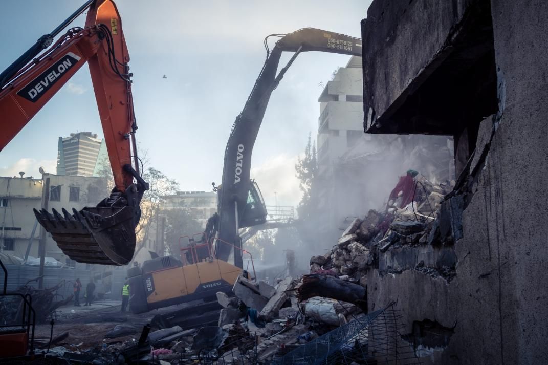 Dos excavadoras demuelen un edificio cubierto de escombros en una zona urbana en construcción, envuelta en humo.