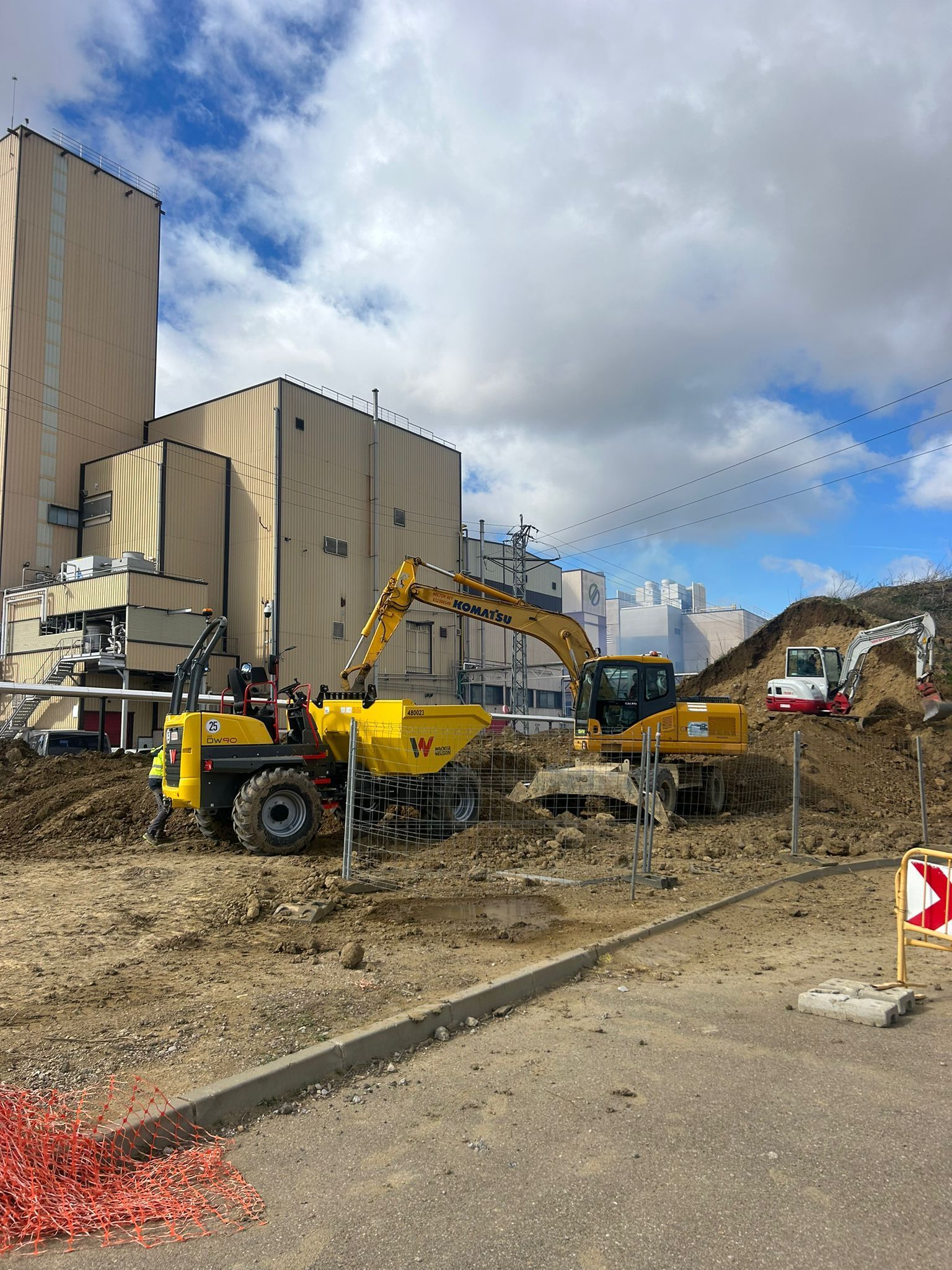 Obra en construcción con excavadora amarilla junto a grandes edificios industriales bajo un cielo nublado.