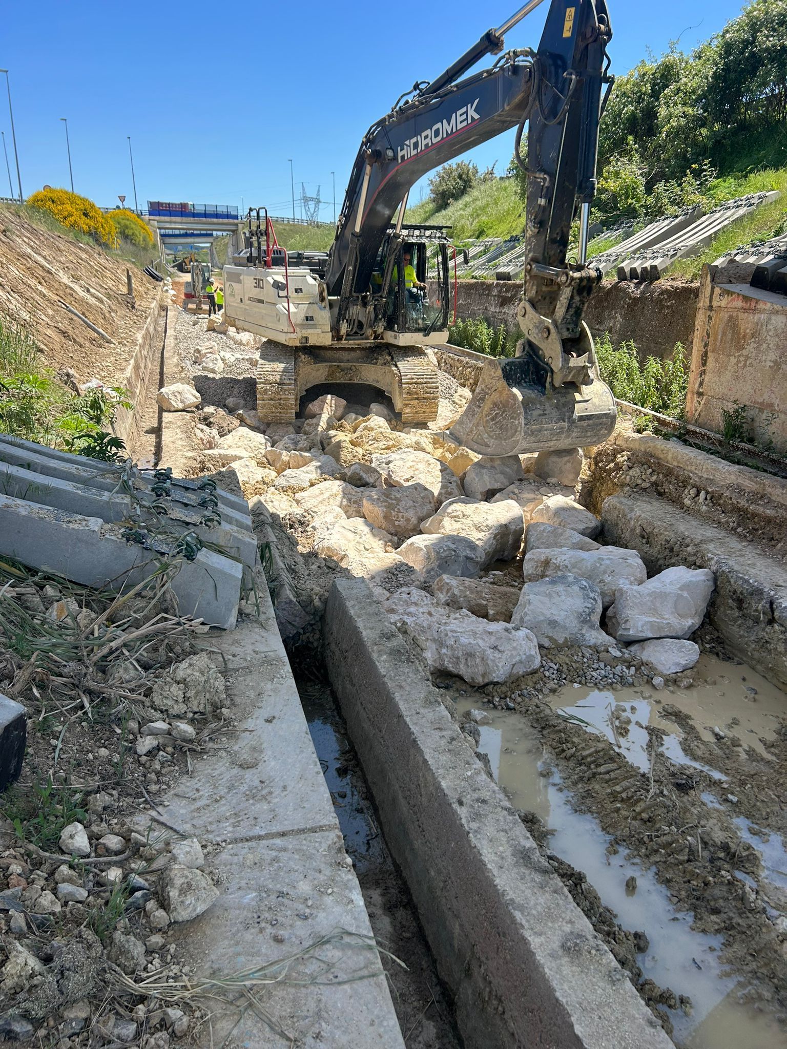 Una excavadora retira escombros en una zanja estrecha junto a una carretera bajo un cielo azul despejado.