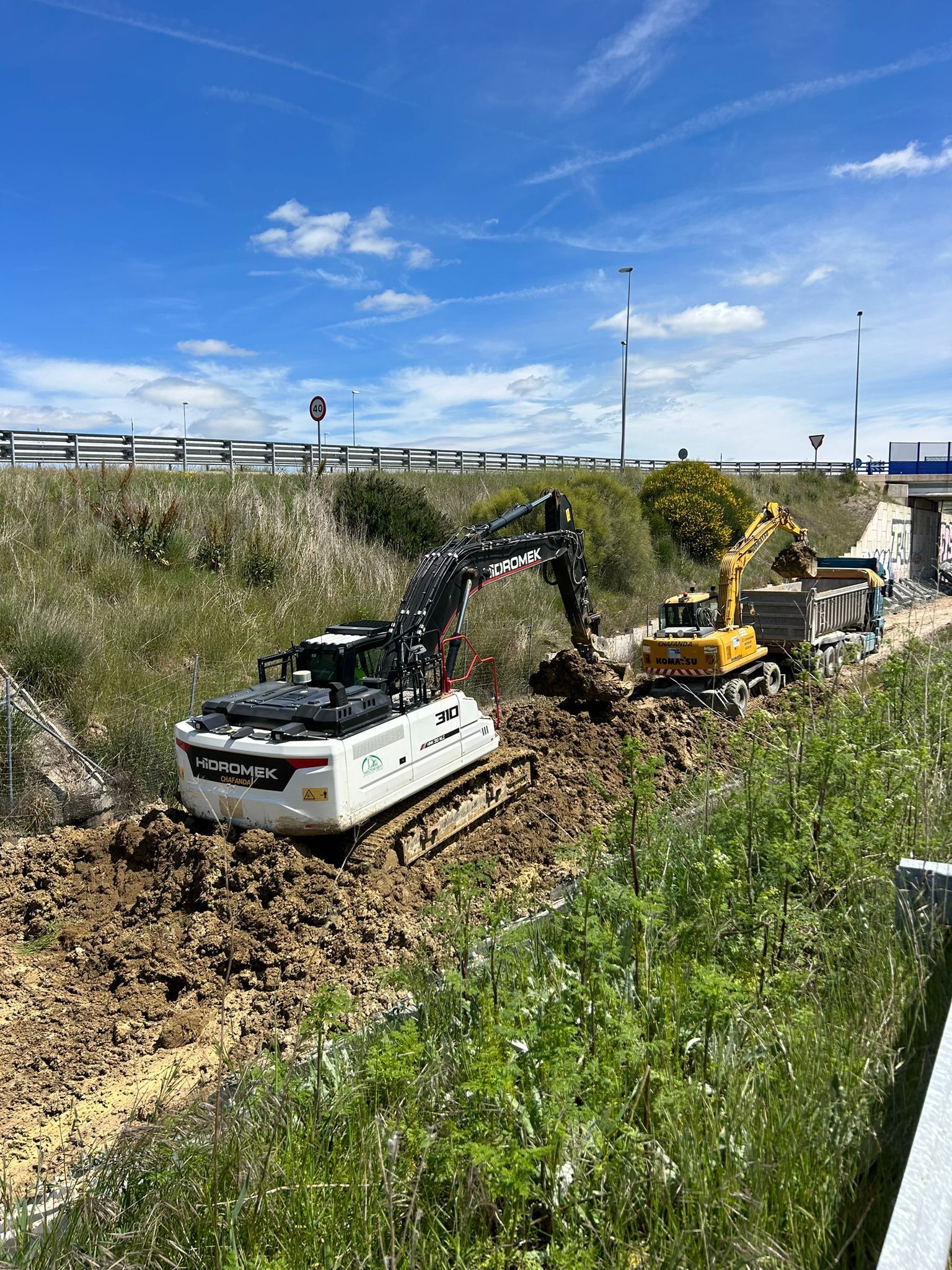 Dos excavadoras cavando una zanja de drenaje fangosa junto a hierba alta bajo un cielo azul.