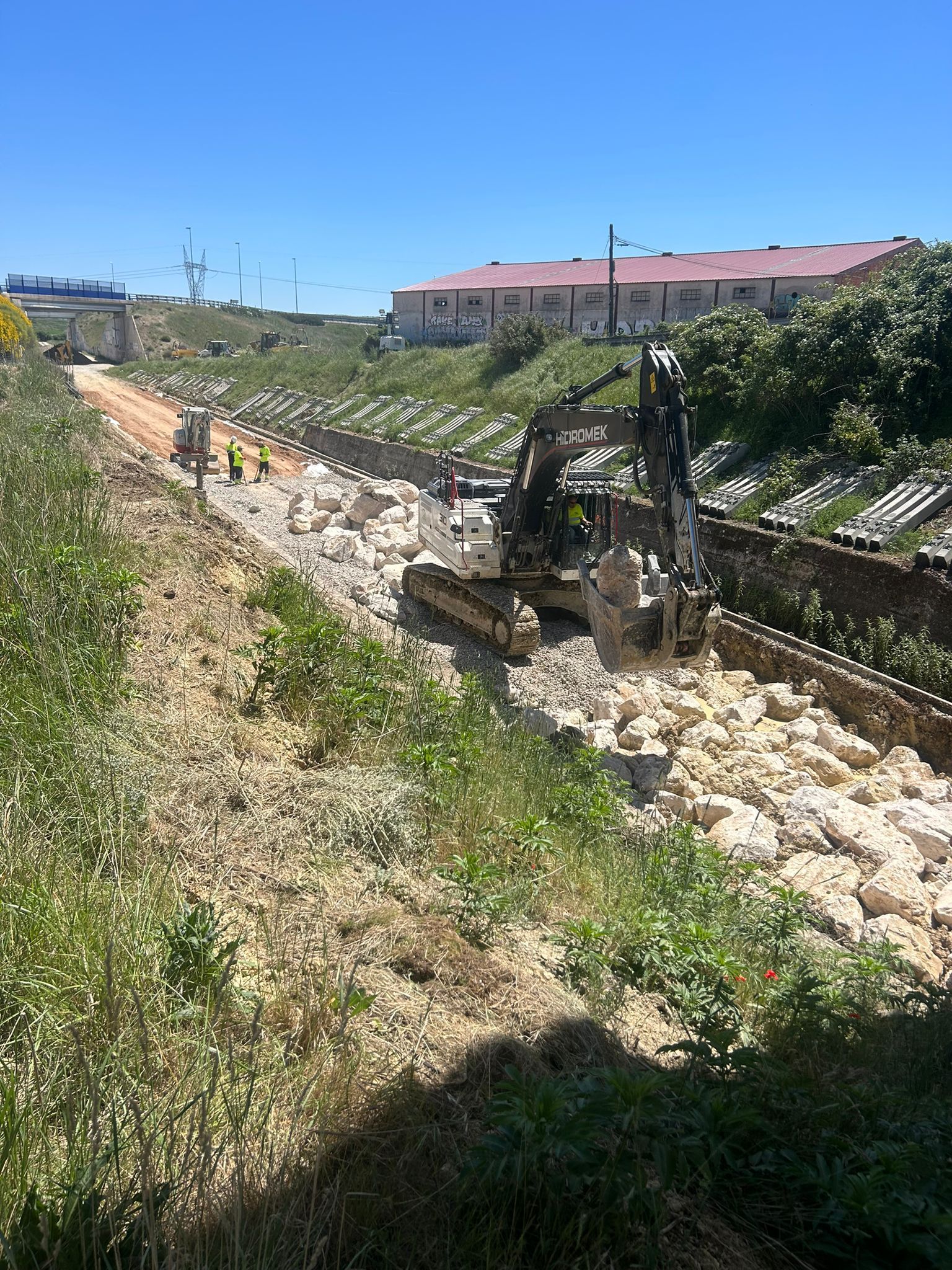 Vía férrea de mantenimiento junto a una ladera con rocas, postes y un edificio de techo rojo bajo un cielo despejado.