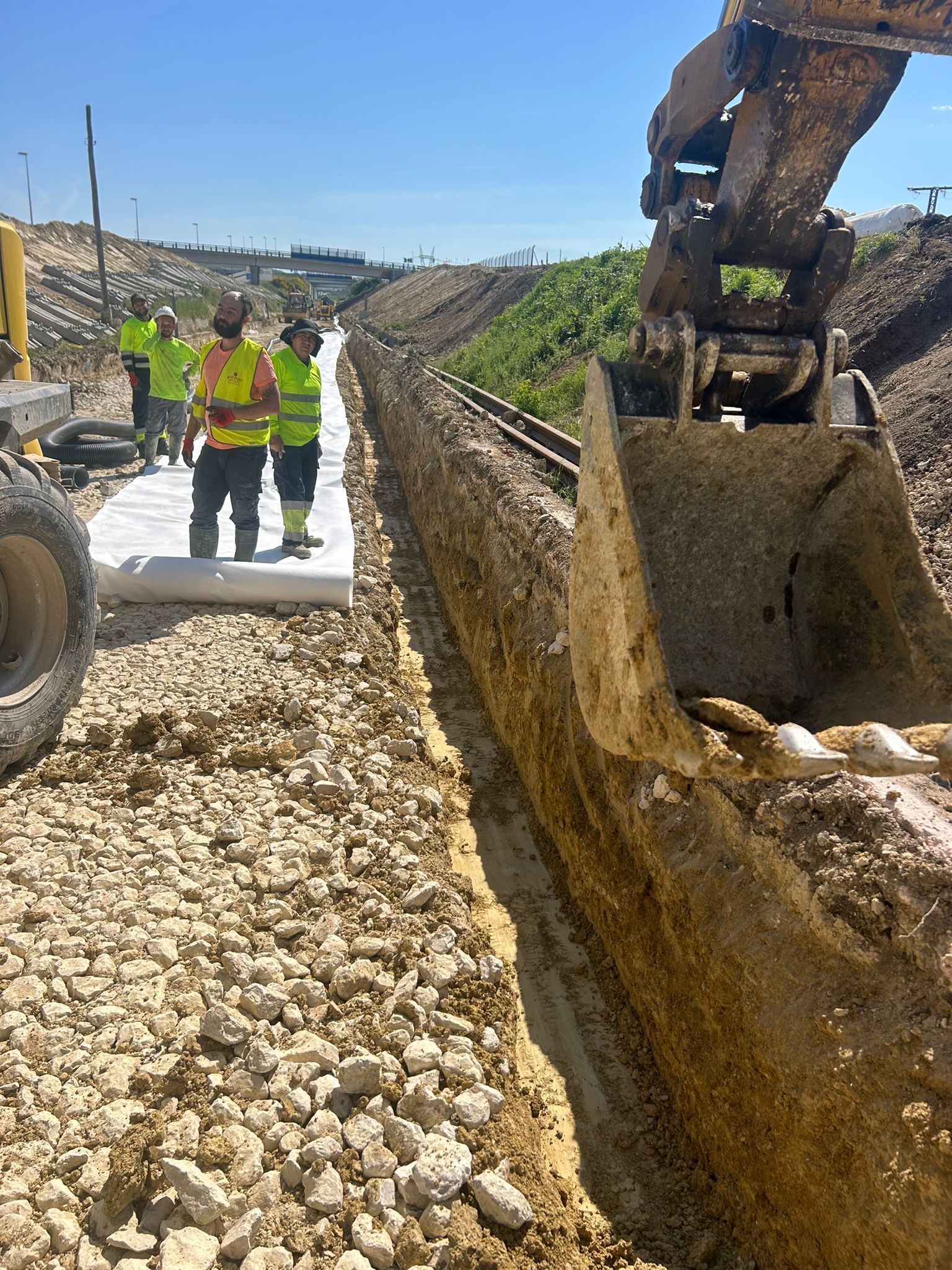 Obreros de la construcción junto a una zanja mientras una excavadora retira grava