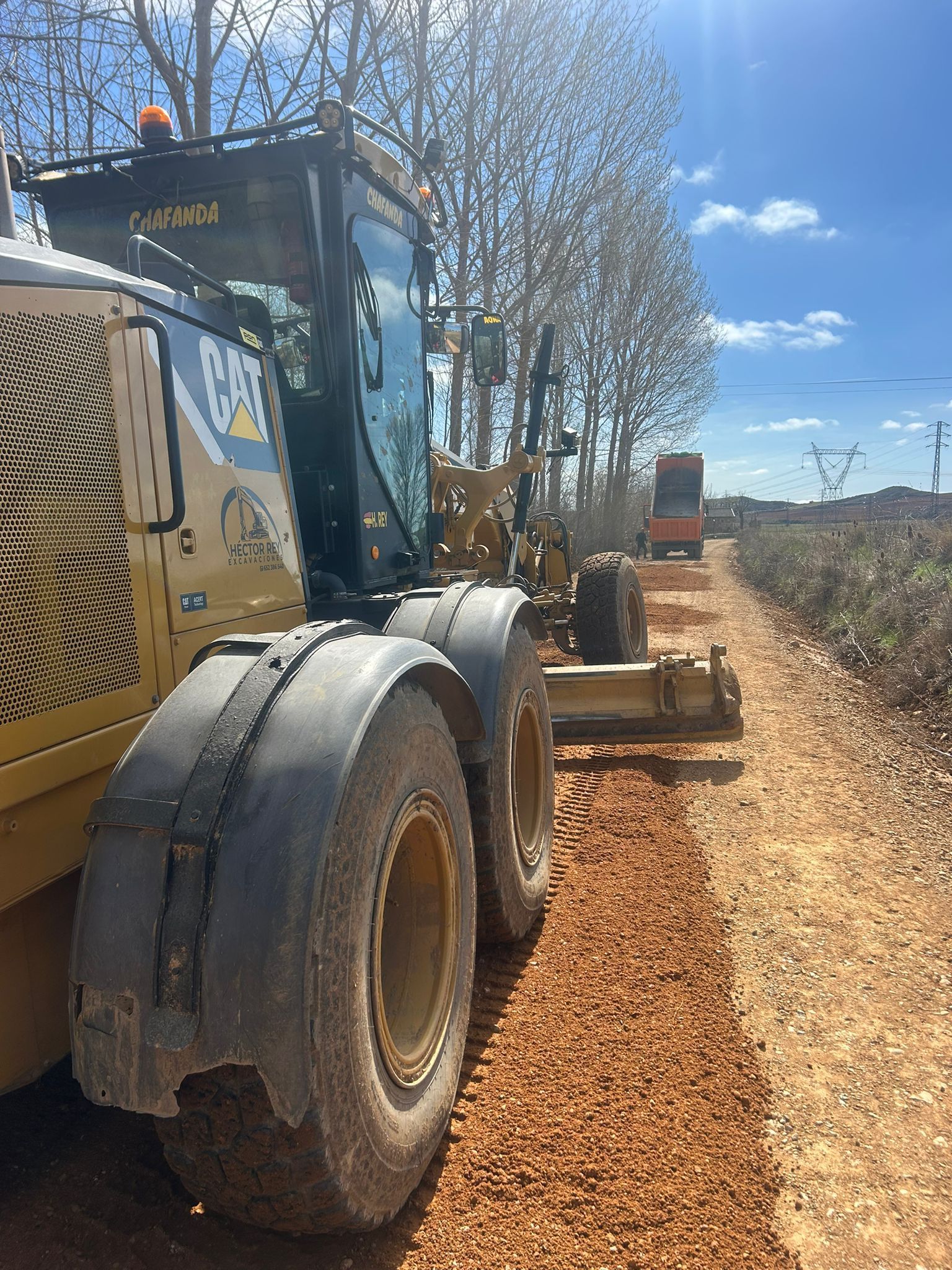 Una motoniveladora trabajando en un camino de tierra junto a árboles bajo un cielo azul.
