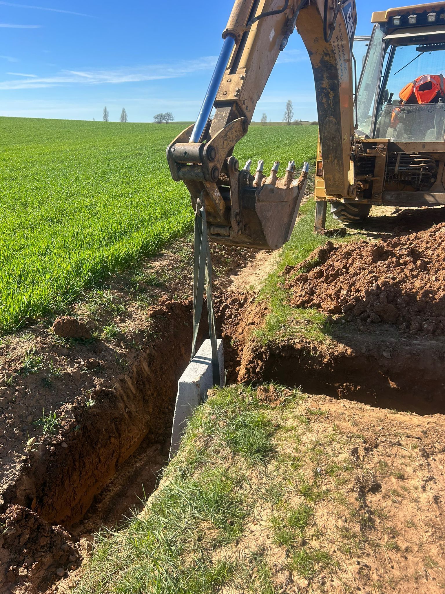 Excavadora abriendo una zanja a lo largo de un campo verde bajo un cielo azul.