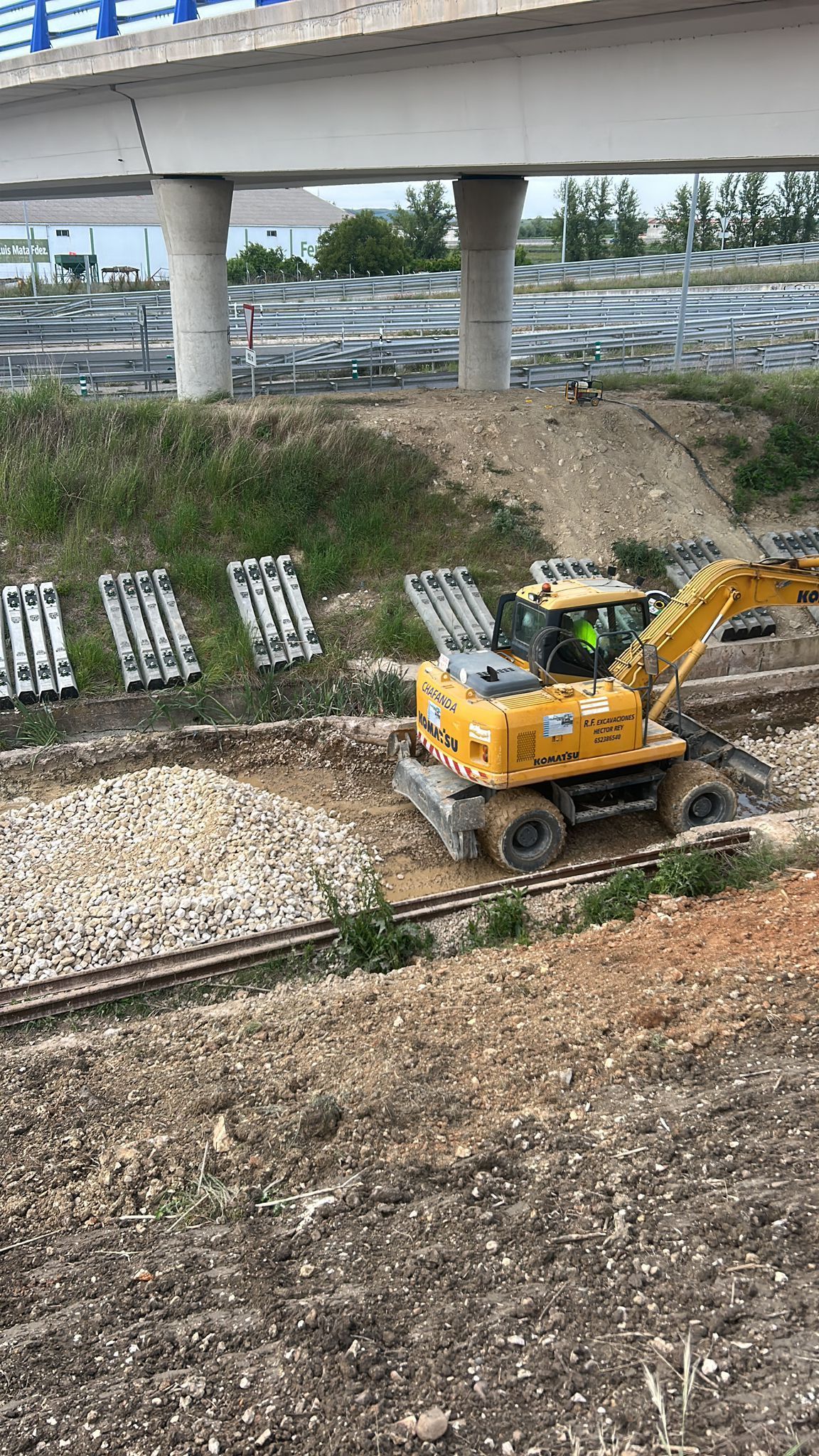 Una excavadora amarilla estacionada sobre grava debajo de un paso elevado de la autopista