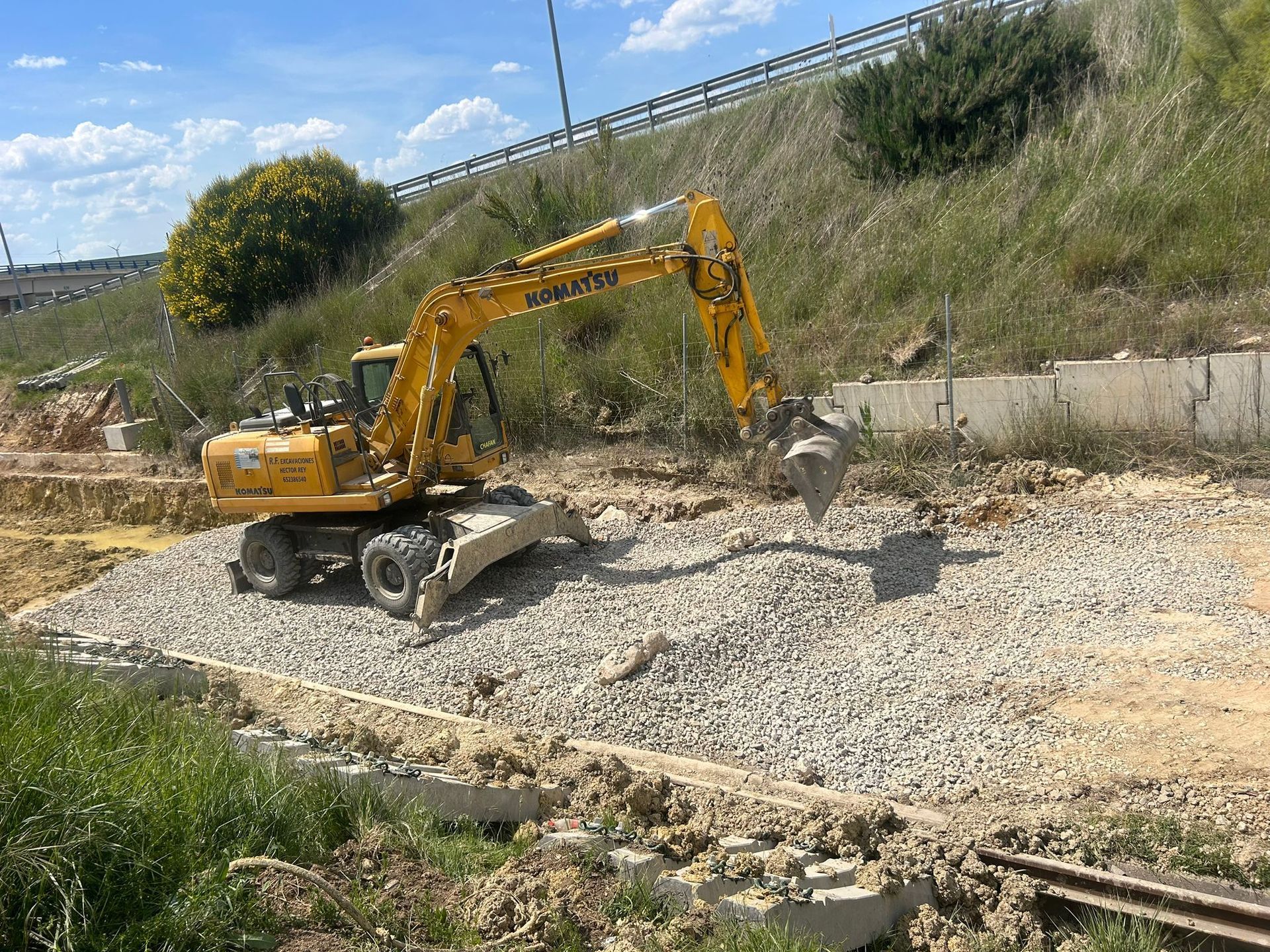 Excavadora amarilla removiendo grava junto a un terraplén al borde de la carretera.