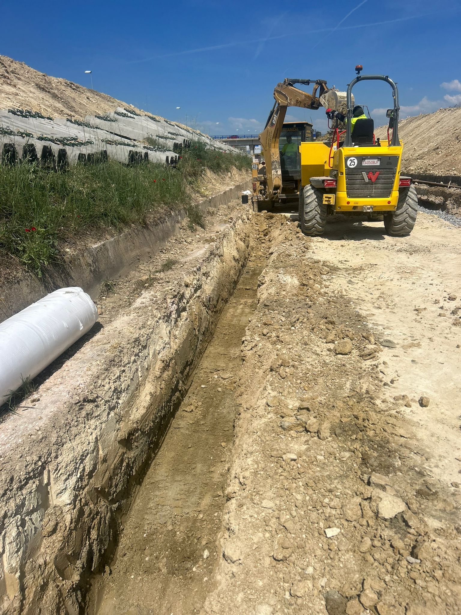 Excavadora amarilla cavando una zanja junto a un camino de tierra en una obra de construcción en una ladera seca.