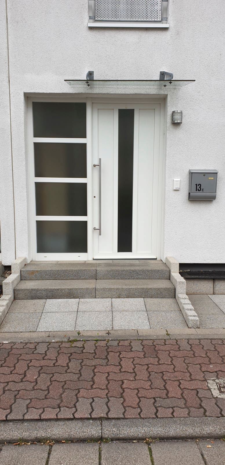 Auf einem gepflasterten Gehweg steht eine weiße Tür mit einem Glasdach darüber.