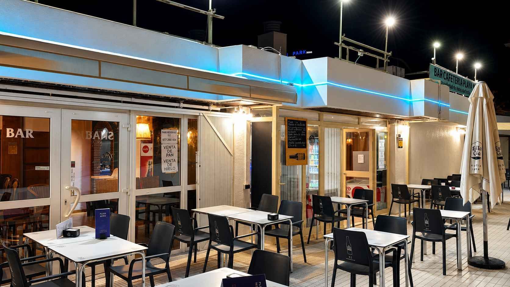 Terraza de una cafetería con mesas al aire libre bajo un toldo iluminado por la noche, con mesas y sillas .