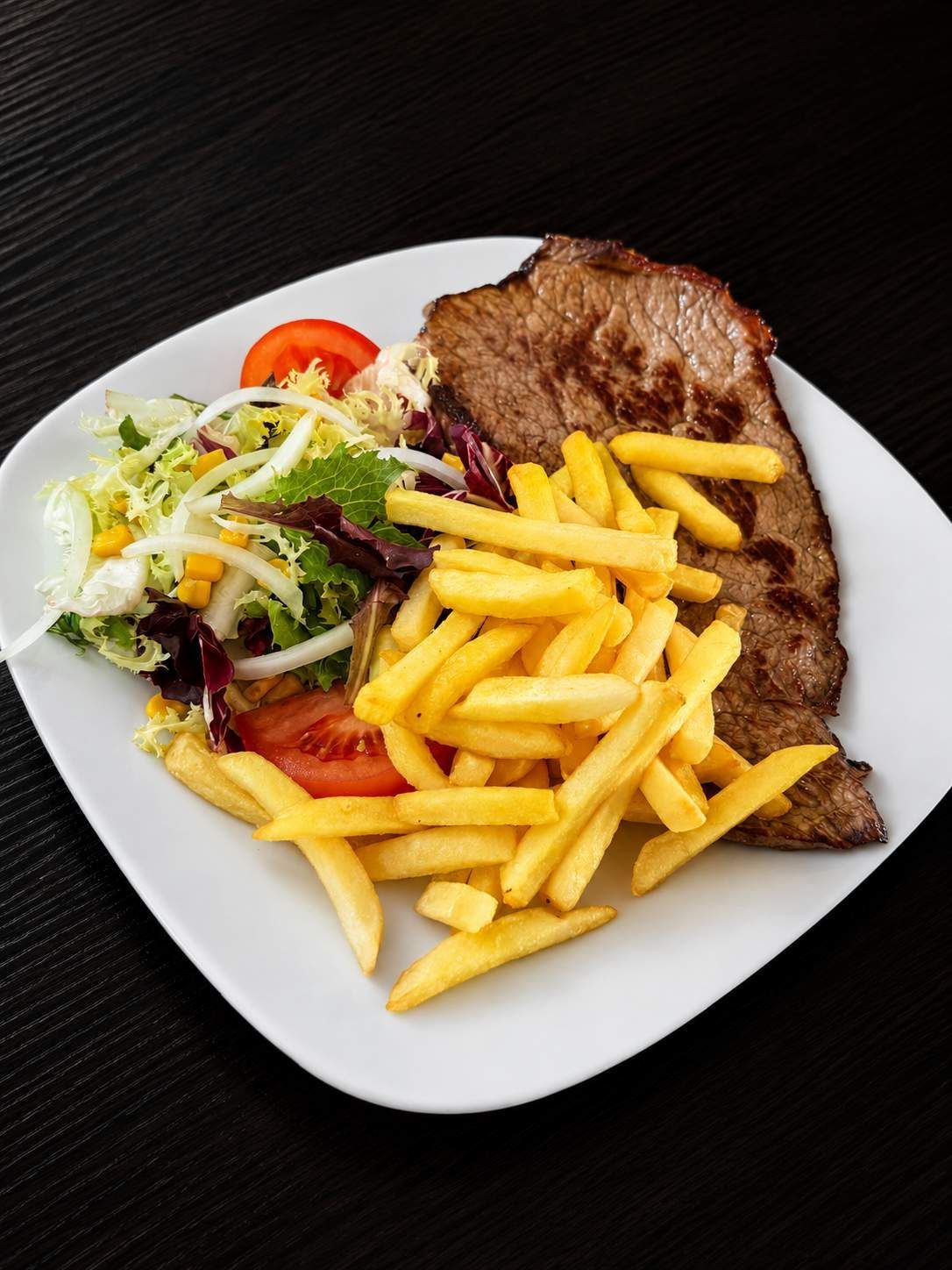 Filete a la parrilla con papas fritas y ensalada en un plato blanco sobre un fondo oscuro.