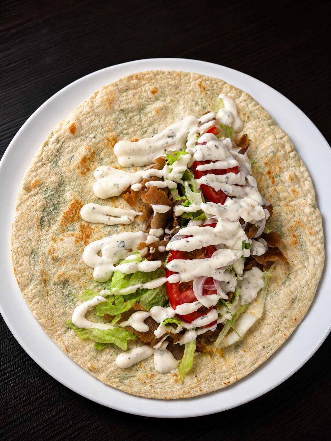 Wrap de pan plano con verduras en rodajas, carne y salsa blanca en un plato blanco.