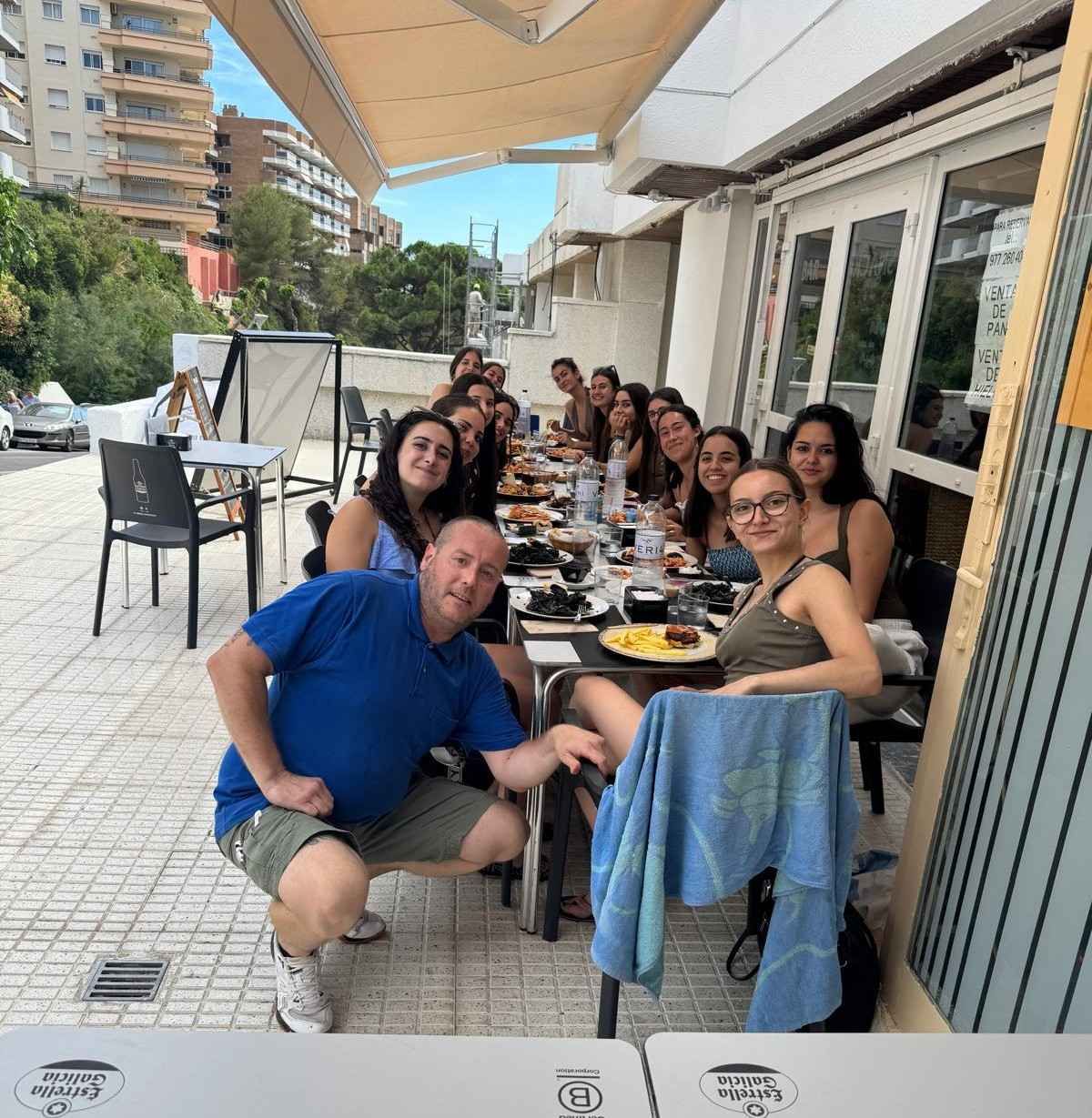 Un grupo numeroso cena al aire libre en una mesa larga en una terraza, sonriendo y posando juntos.