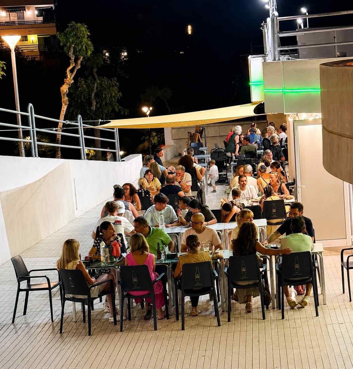 Gente sentada en mesas en la terraza de un restaurante nocturno al aire libre con luces brillantes y paredes blancas.