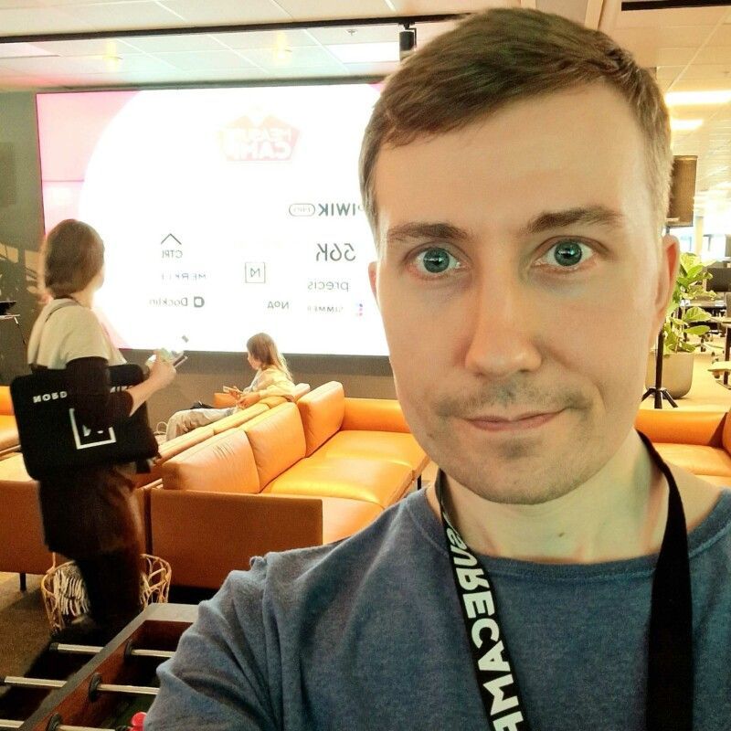 Selfie olohuoneessa, jossa on oransseja sohvia, kirkas seinätaulu ja taustalla ihmisiä