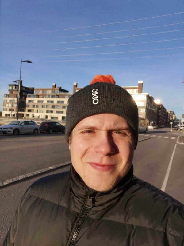 Mustapipoinen ja -takkinen mies seisoo aurinkoisella kaupungin kadulla, takanaan kerrostalot