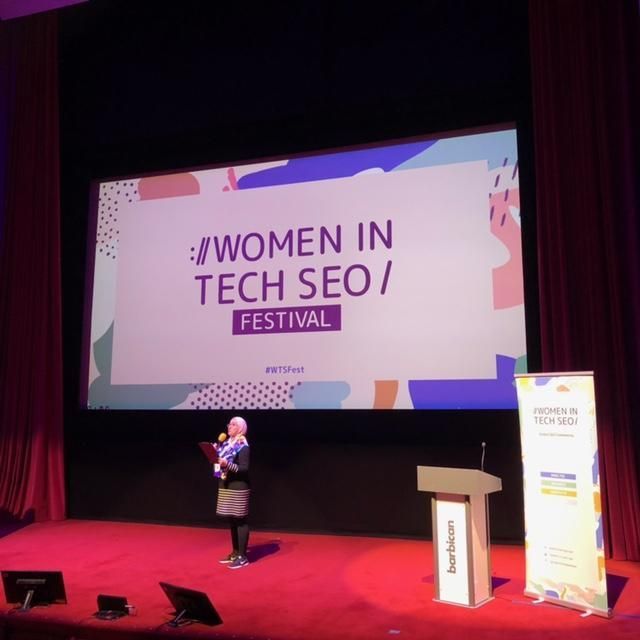 Puhuja punaisella lavalla Women in Tech SEO -festivaaleilla suuren esitysnäytön ja puhujakorokkeen kanssa.