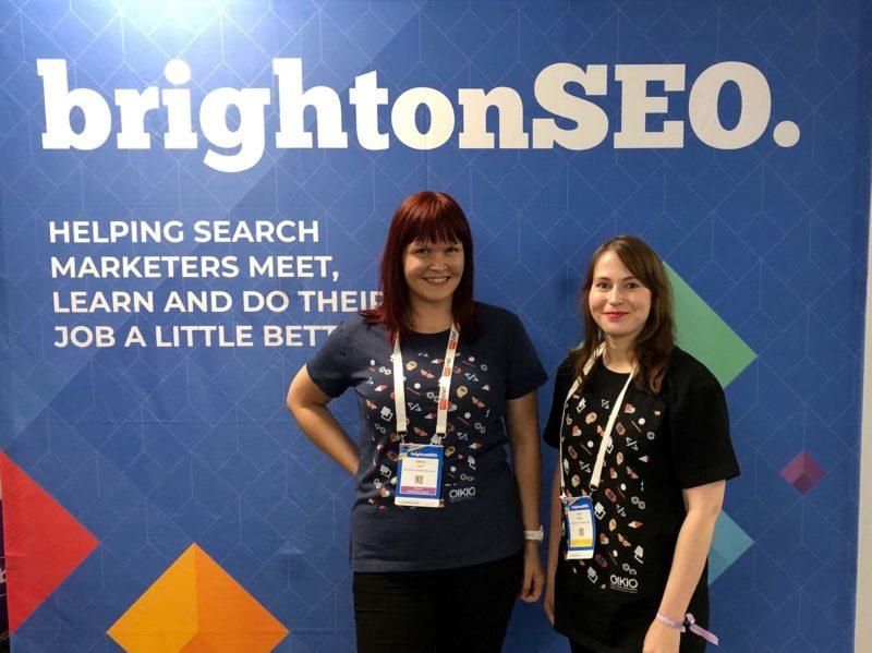 Kaksi ihmistä seisoo BrightonSEO-tapahtuman taustalla hymyillen ja konferenssikortit päässään.