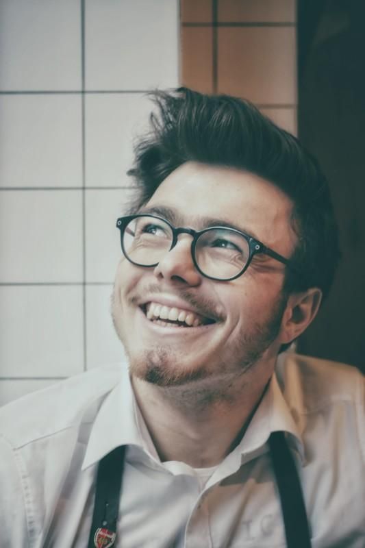 Hymyilevä, ylöspäin katsova henkilö lasit päässään, laatoitettujen seinien edessä