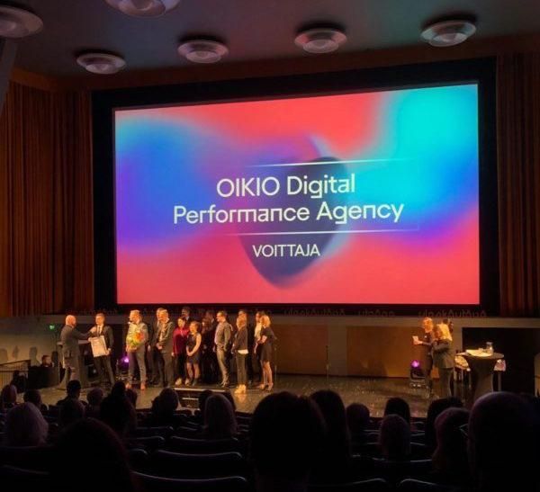 Ihmisiä lavalla suuren valkokankaan alla lukemassa tekstiä ”OIKIO Digital Performance Agency” teatterissa