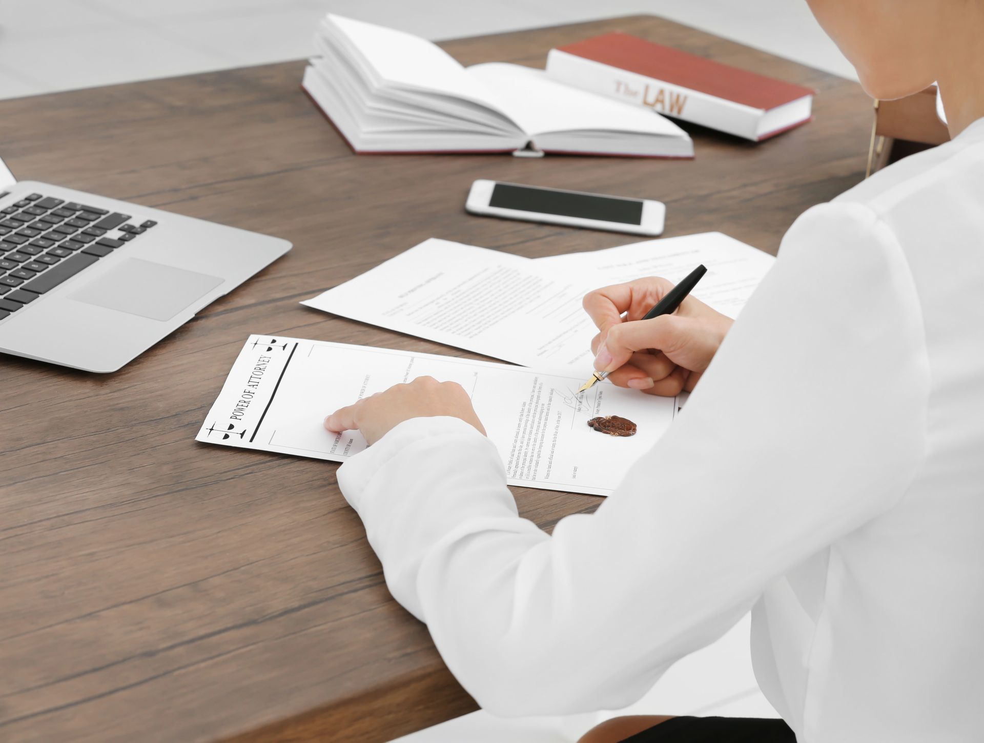 Une personne en chemise blanche rédige des documents à un bureau en bois, avec un ordinateur portable, des livres et un smartphone.