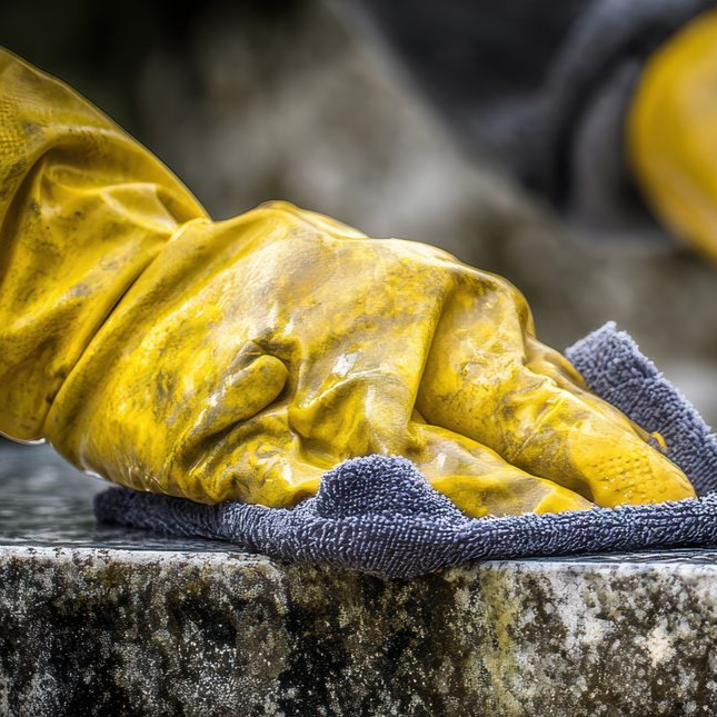 Une main gantée de caoutchouc jaune utilise un chiffon gris pour frotter une surface de pierre texturée.