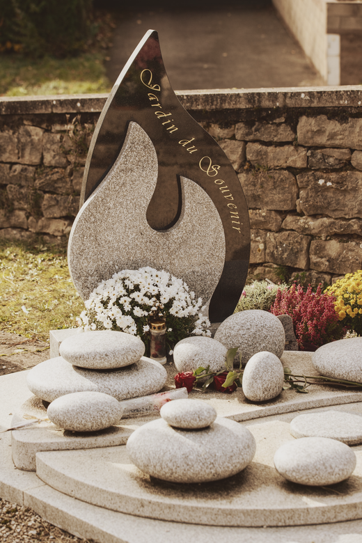 Une pierre tombale moderne en granit ornée d'un monument en forme de flamme avec des inscriptions dorées, entourée d'éléments en pierre aux formes arrondies.