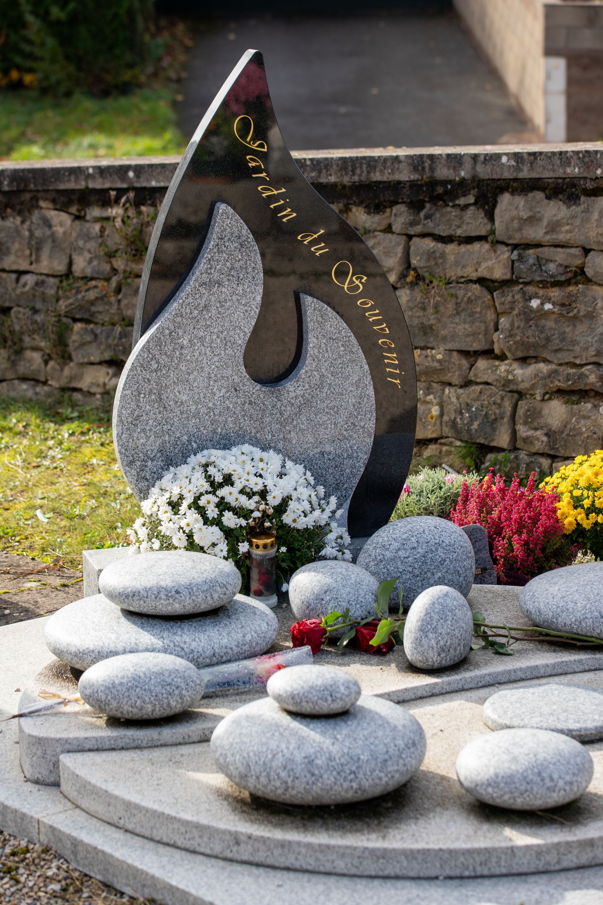 Une pierre tombale en granit ornée d'une sculpture en forme de flamme, composée de galets, de fleurs blanches et de plantes sur un socle à degrés.