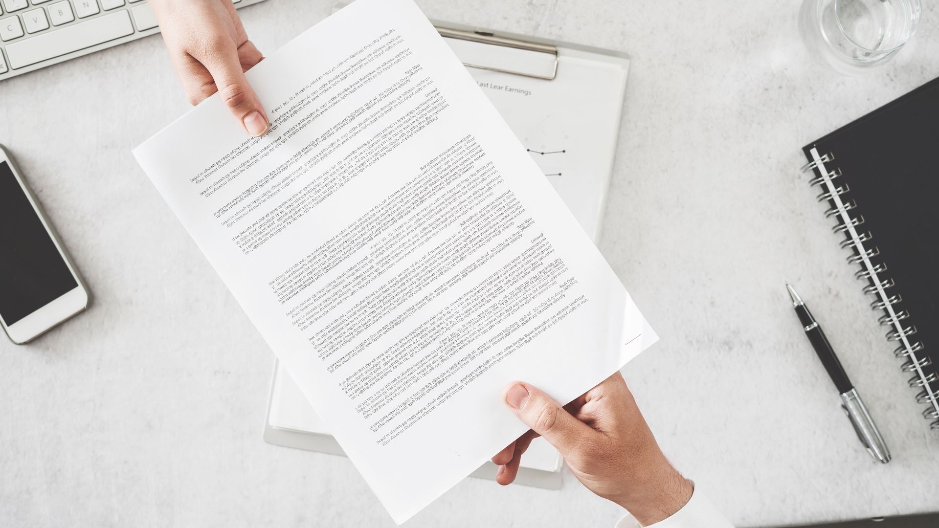 Deux personnes tenant un document papier au-dessus d'un bureau, avec un ordinateur portable, un smartphone, un stylo et un carnet.