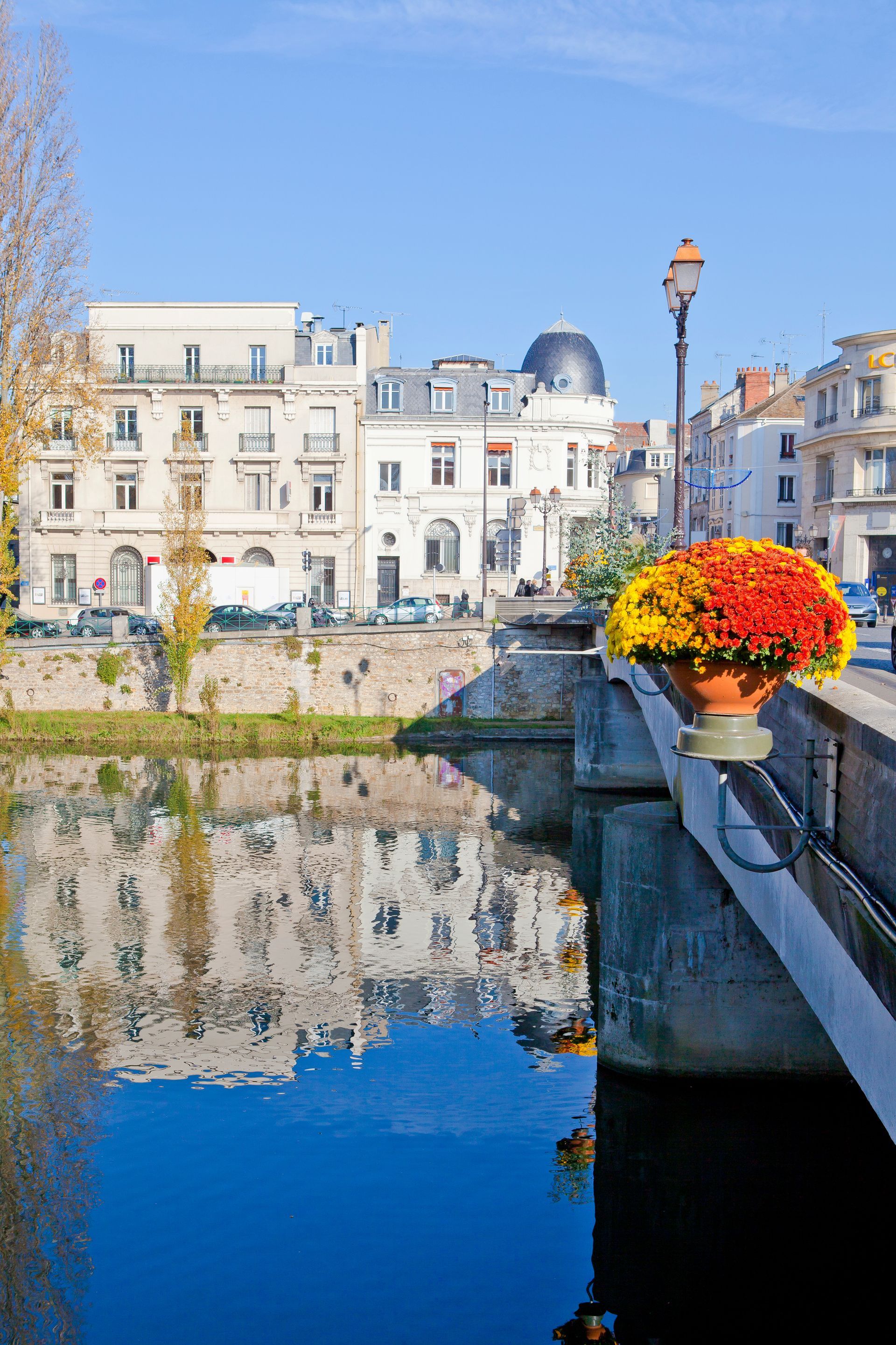 Des bâtiments et leur reflet dans une rivière calme, vus depuis un pont de pierre orné d'un pot aux couleurs éclatantes de fleurs d'automne.
