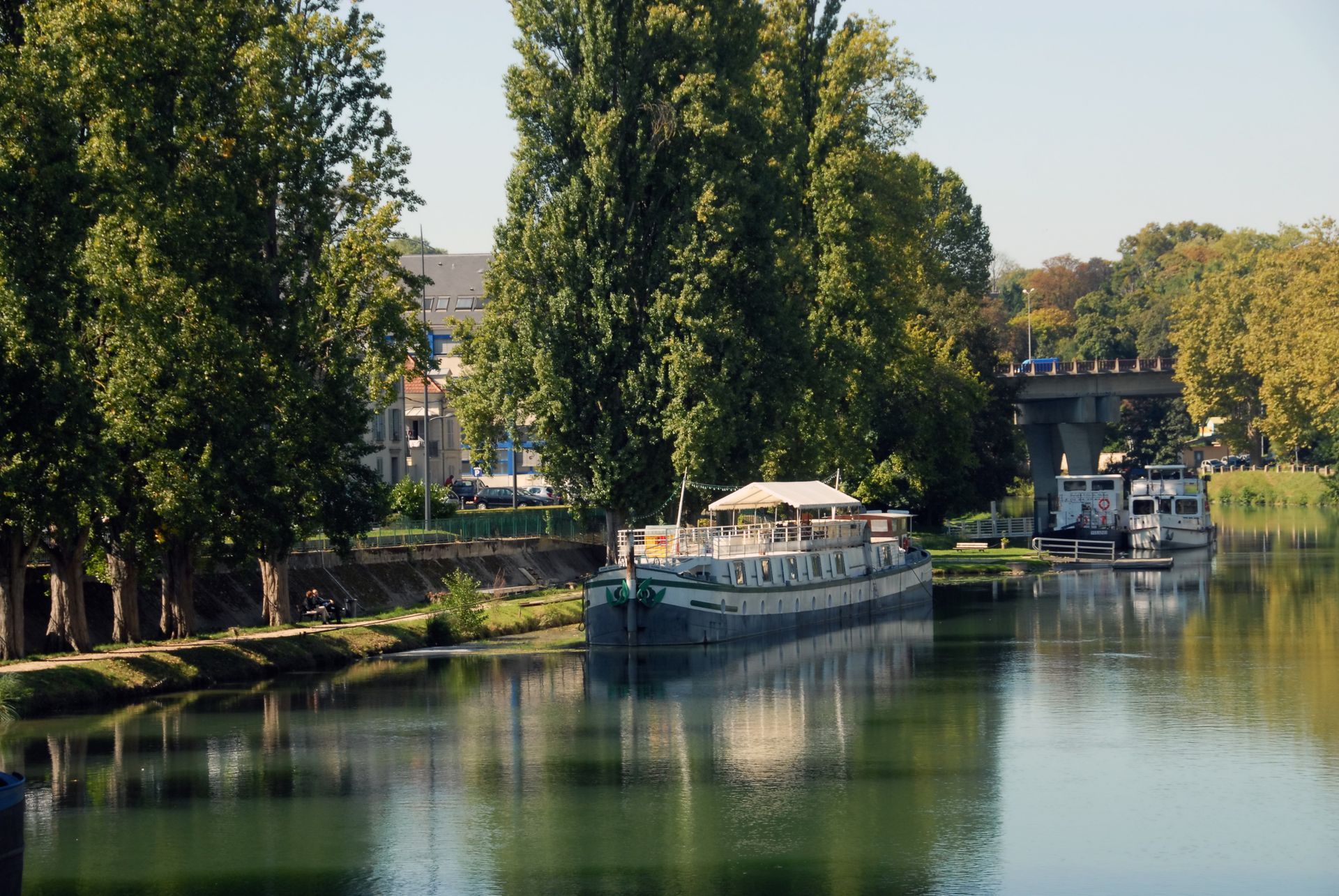 Un canal étroit bordé de grands arbres, avec une grande péniche résidentielle blanche amarrée dans les eaux calmes et miroitantes.
