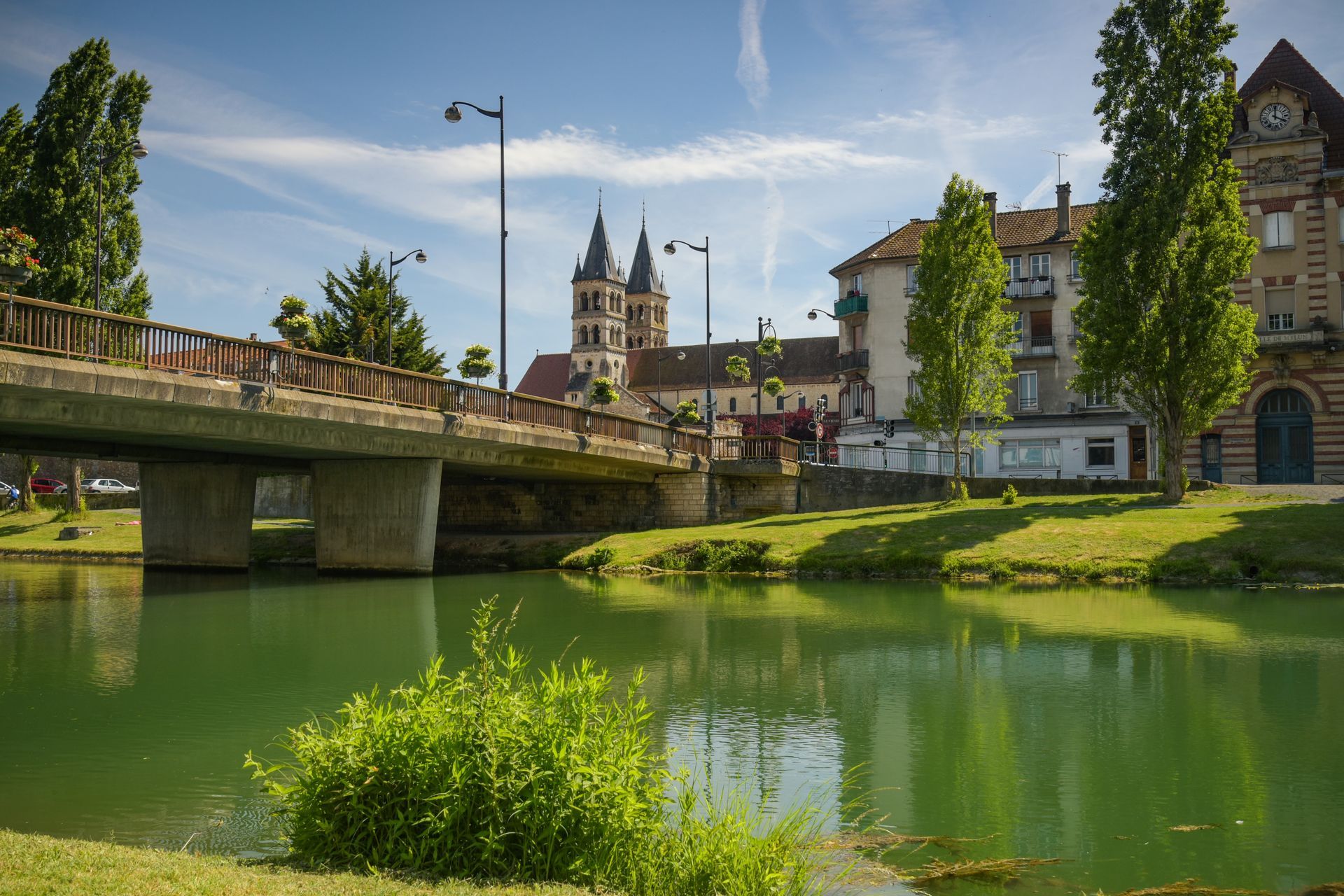 Une rivière coule sous un pont, avec une église à deux tours et des bâtiments en arrière-plan, sous un ciel bleu.
