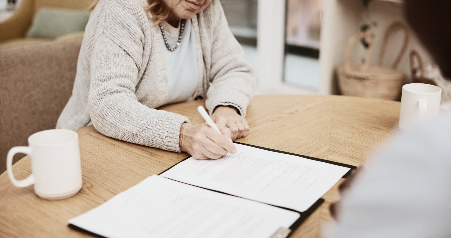 Une personne assise à une table en bois signe un document tandis qu'une tasse blanche se trouve à proximité.