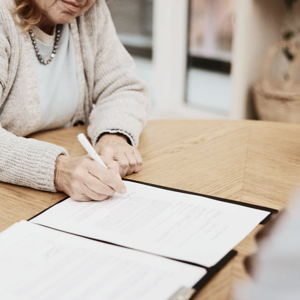 Une personne vêtue d'un cardigan clair est assise à une table en bois, en train d'écrire sur un document rangé dans un dossier noir.
