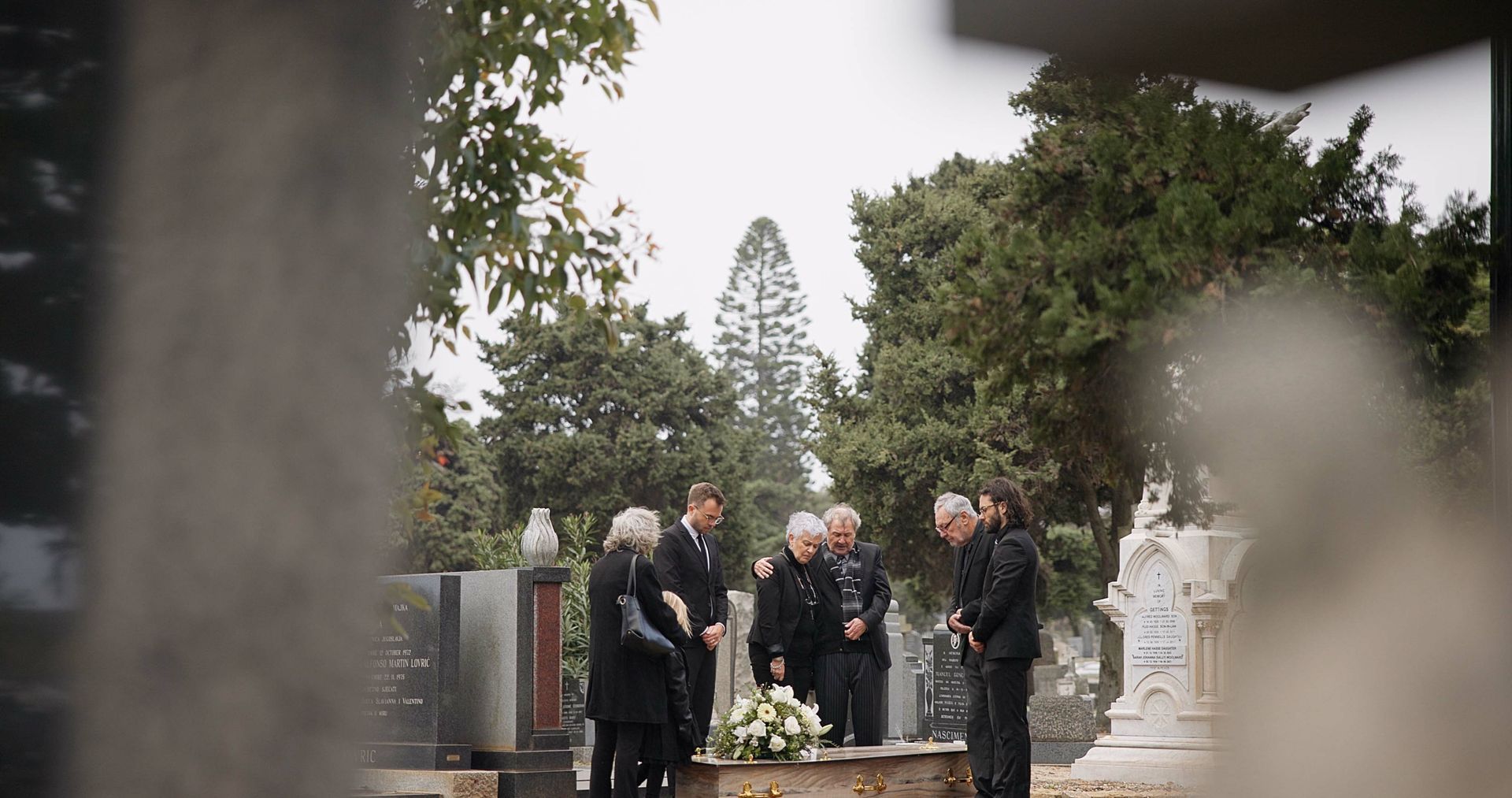 Un petit groupe de personnes vêtues de noir se tient solennellement autour d'une composition florale dans un cimetière.